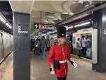 A man wearing a British soldier outfit at 2nd Ave Station walking on the platform between the R179 consist F train and the Arnine consist making up the Holiday Train